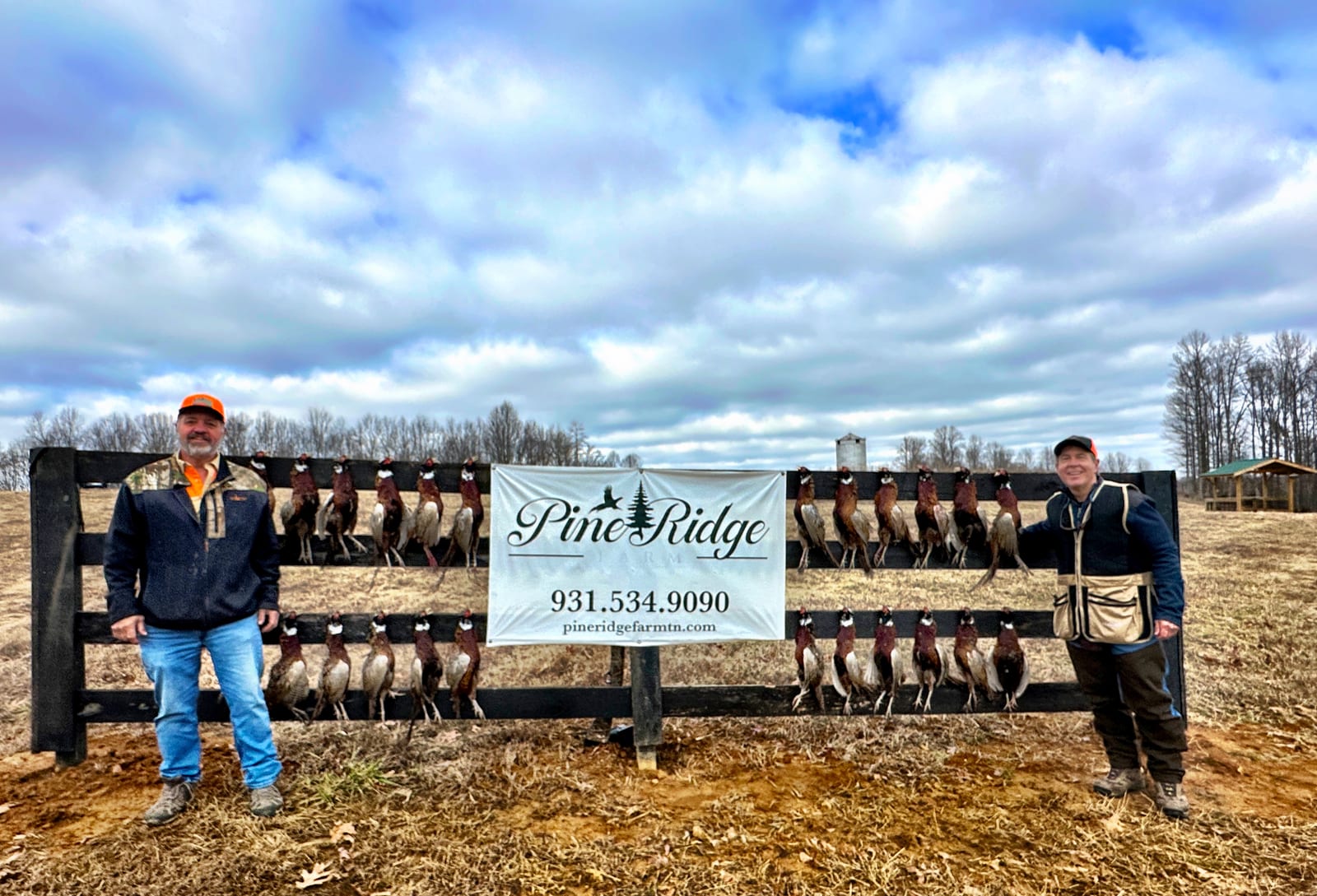 Two men pose with harvested Ring-necked Pheasants hanging on a fence with a Pine Ridge Farms banner.