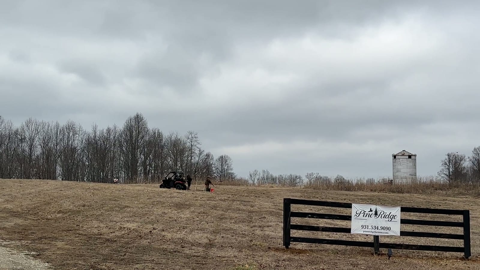 A wide field with bare trees, a fence with a 'Pine Ridge' sign, and a vehicle with a person in the distance under a cloudy sky.
