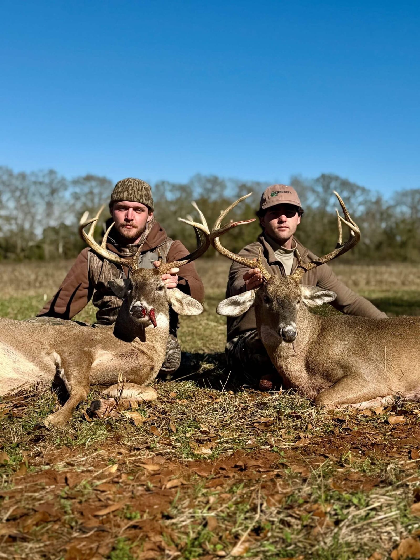 Two hunters pose with two harvested whitetail deer in a field under a clear blue sky.