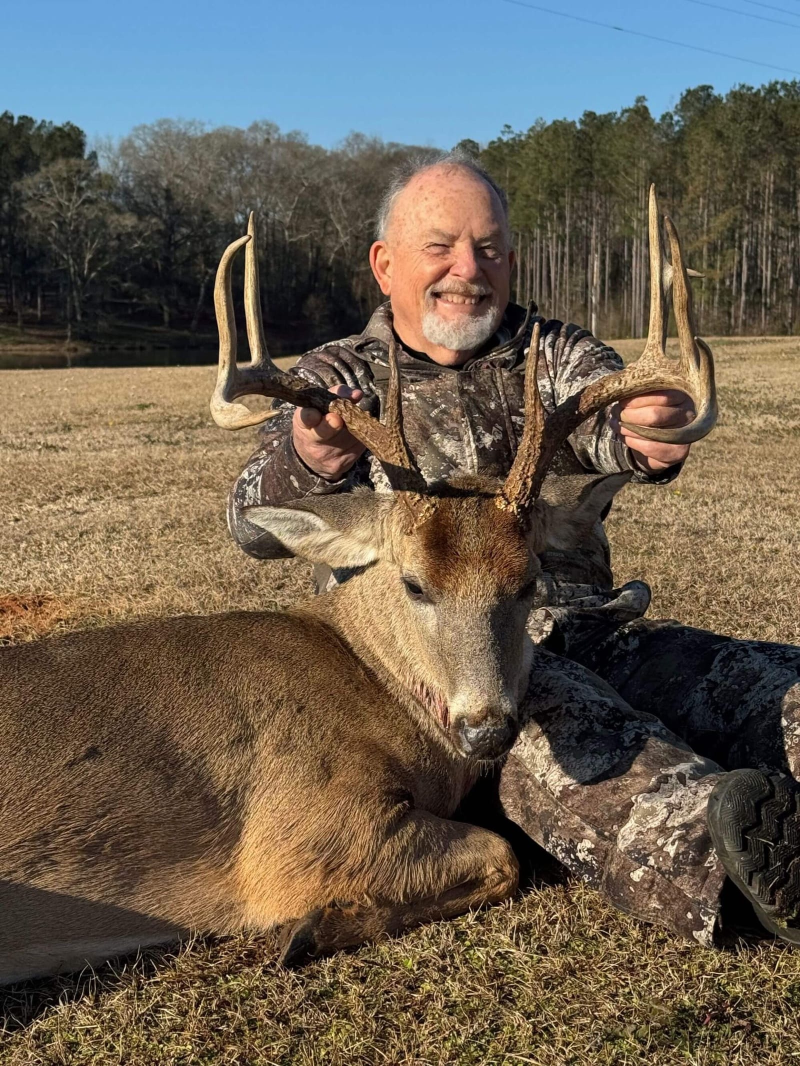 A smiling hunter in camouflage poses with a harvested whitetail deer in a grassy field.