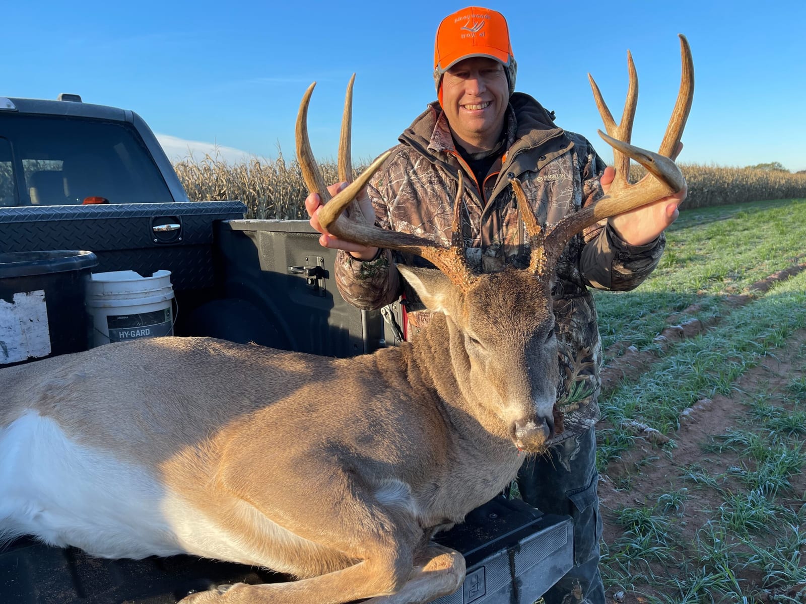 A hunter in camouflage and an orange hat poses with a harvested whitetail deer in the back of a truck.