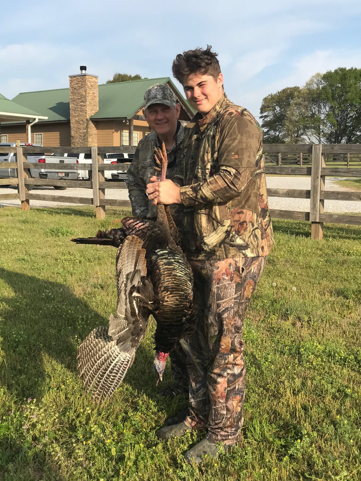Two men in camouflage pose with a harvested wild turkey in a grassy area with a building and fence in the background.