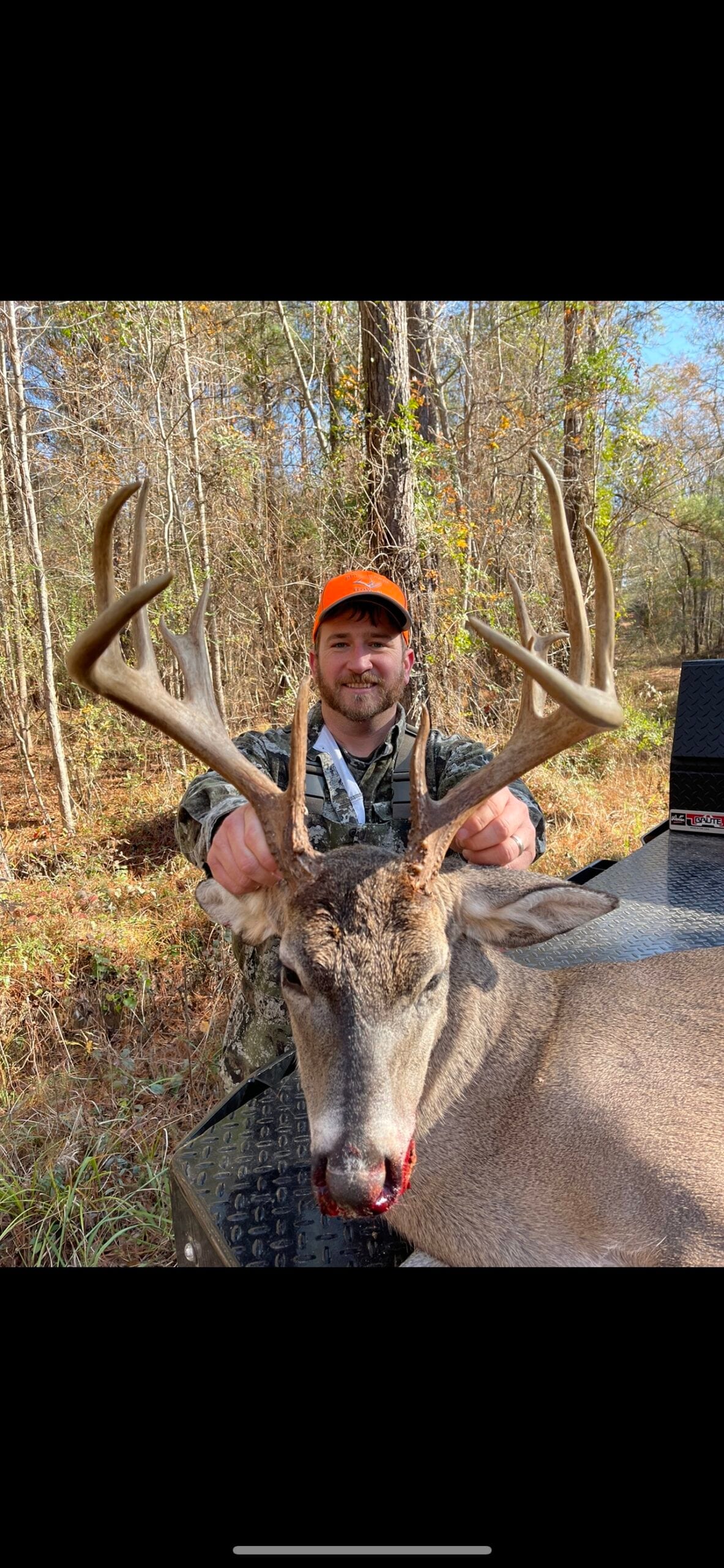 A man in an orange hat poses with a harvested whitetail deer in a wooded area.