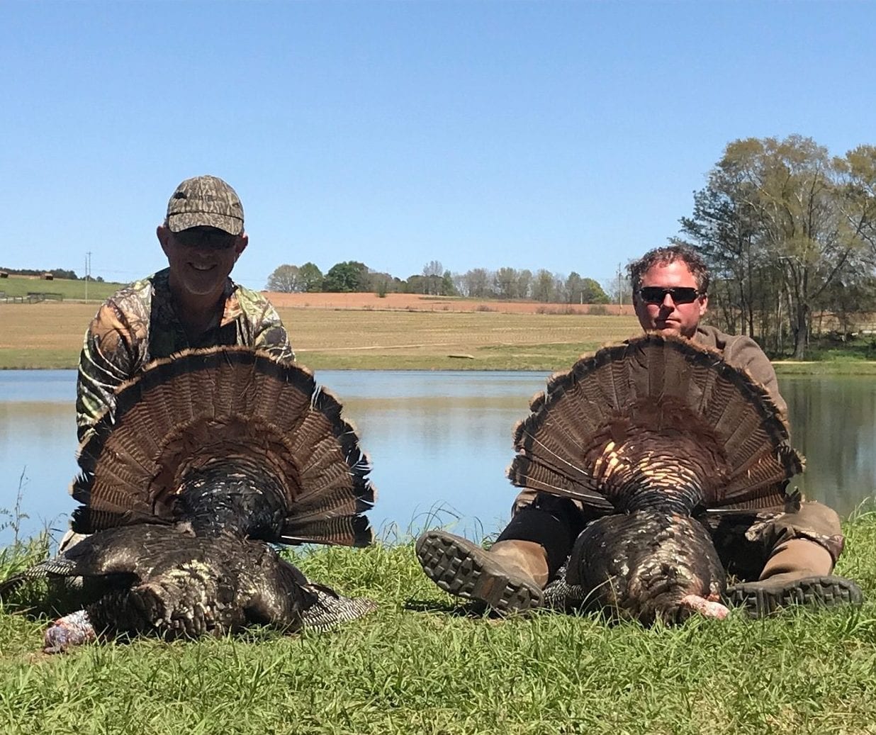 Two hunters pose with two harvested wild turkeys by a pond.