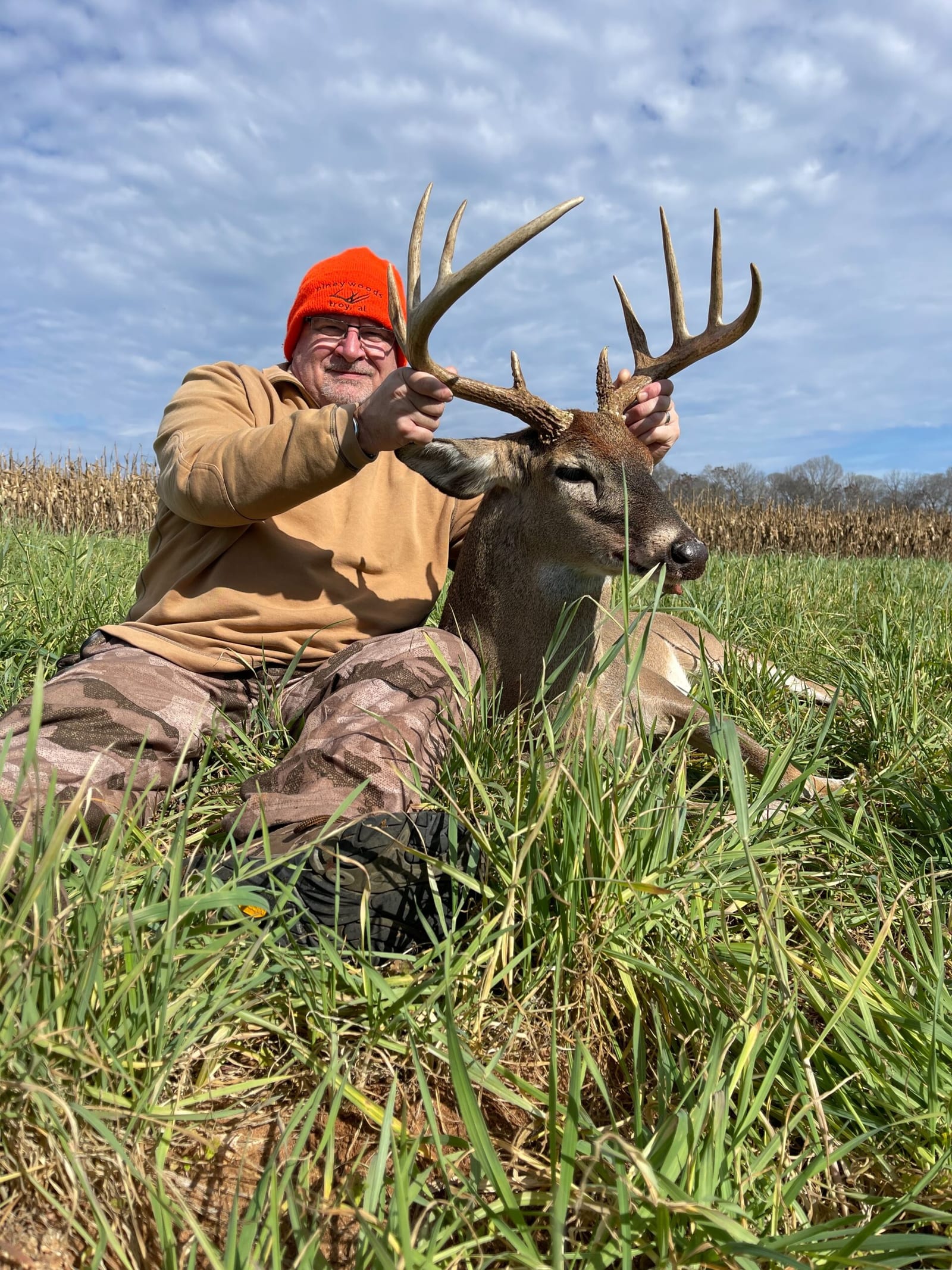 A hunter in an orange hat poses with a harvested whitetail deer in a grassy field.
