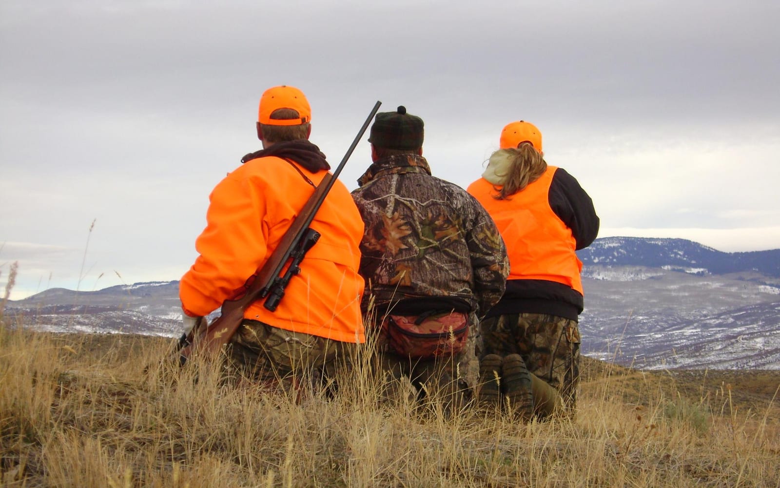 Three hunters in orange and camouflage gear are kneeling in a grassy field, looking out at a distant, snow-dusted mountain range under a cloudy sky.