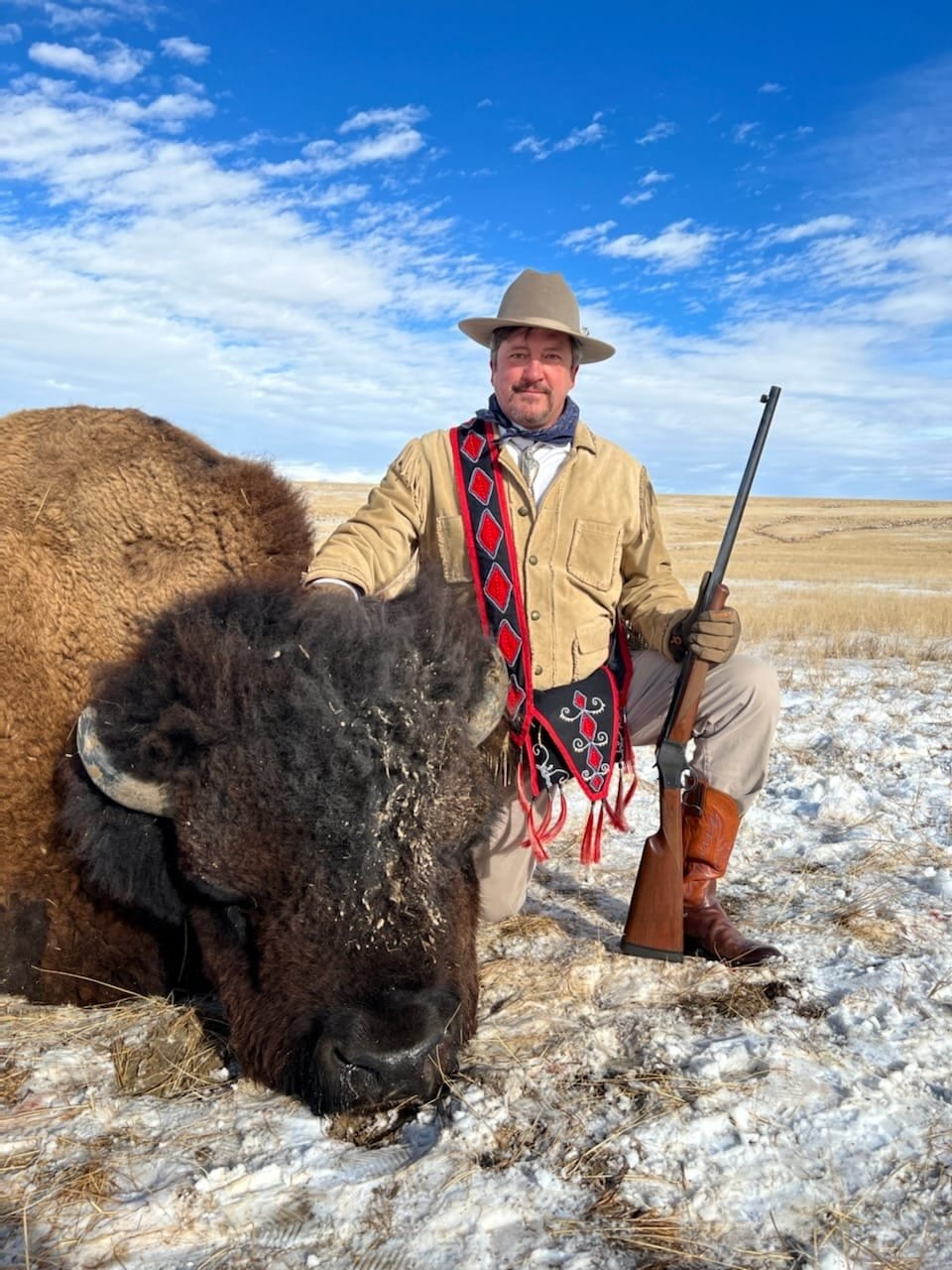 A hunter kneels next to a harvested bison in a snowy field under a blue sky.
