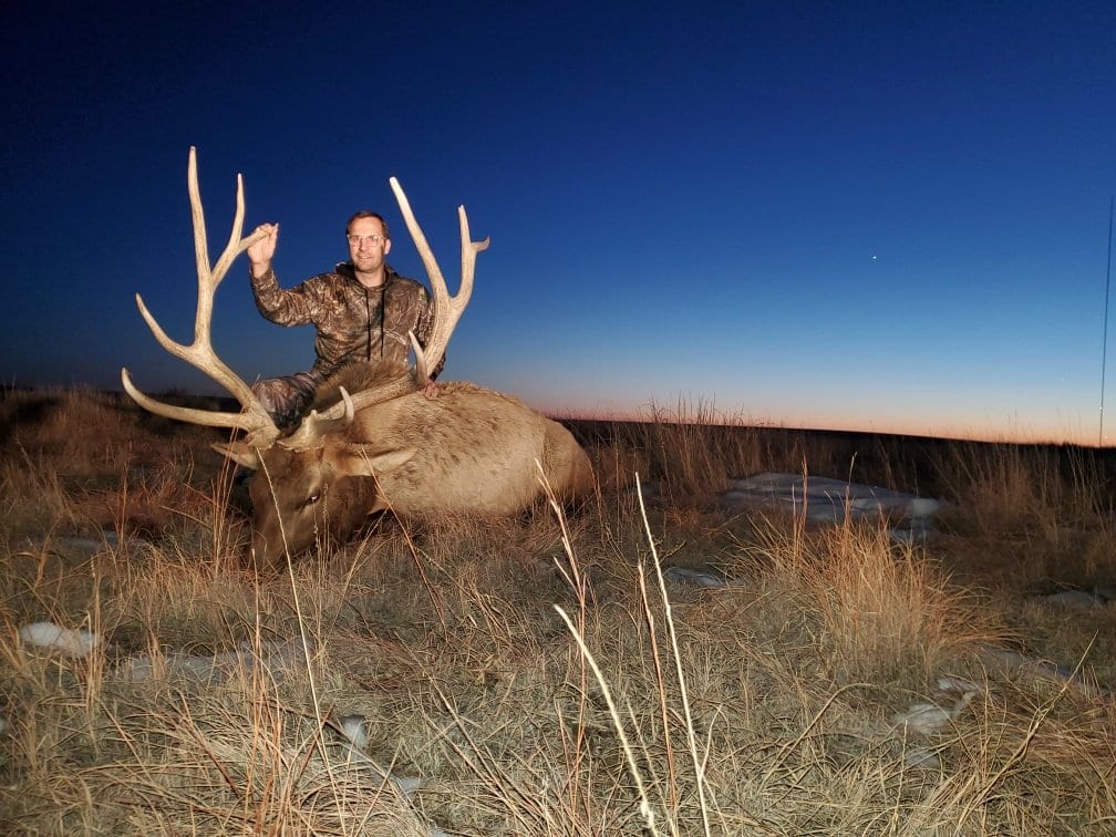 A hunter in camouflage sits on a harvested elk in a field at twilight.