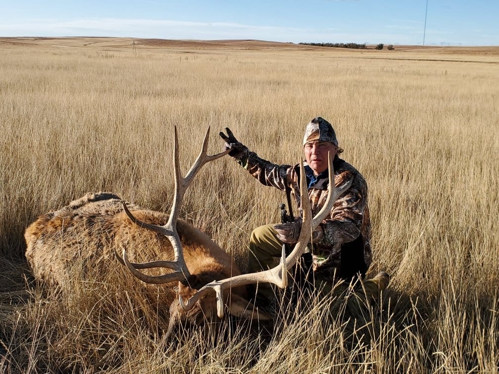 A hunter kneels next to a harvested elk in a grassy field.