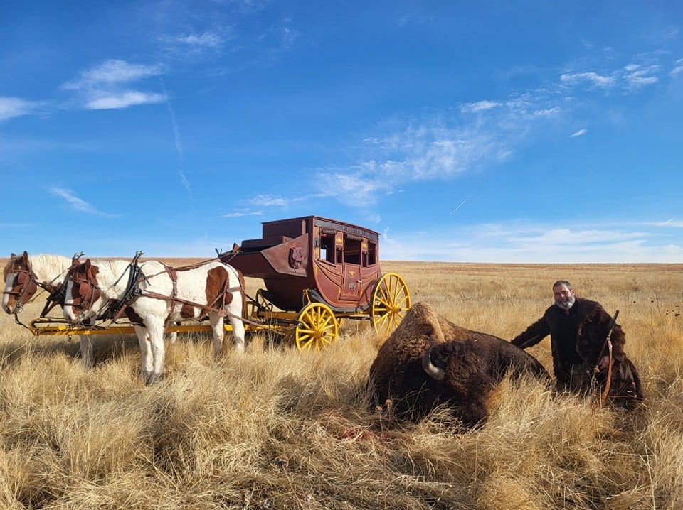 A hunter poses with a harvested bison next to a horse-drawn carriage in a grassy field under a blue sky.