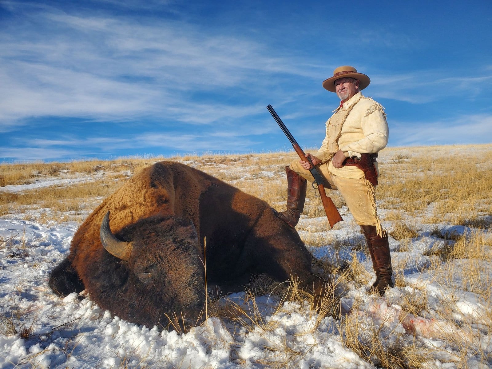 A hunter in a hat and traditional clothing poses with a harvested bison in a snowy, grassy field under a blue sky.