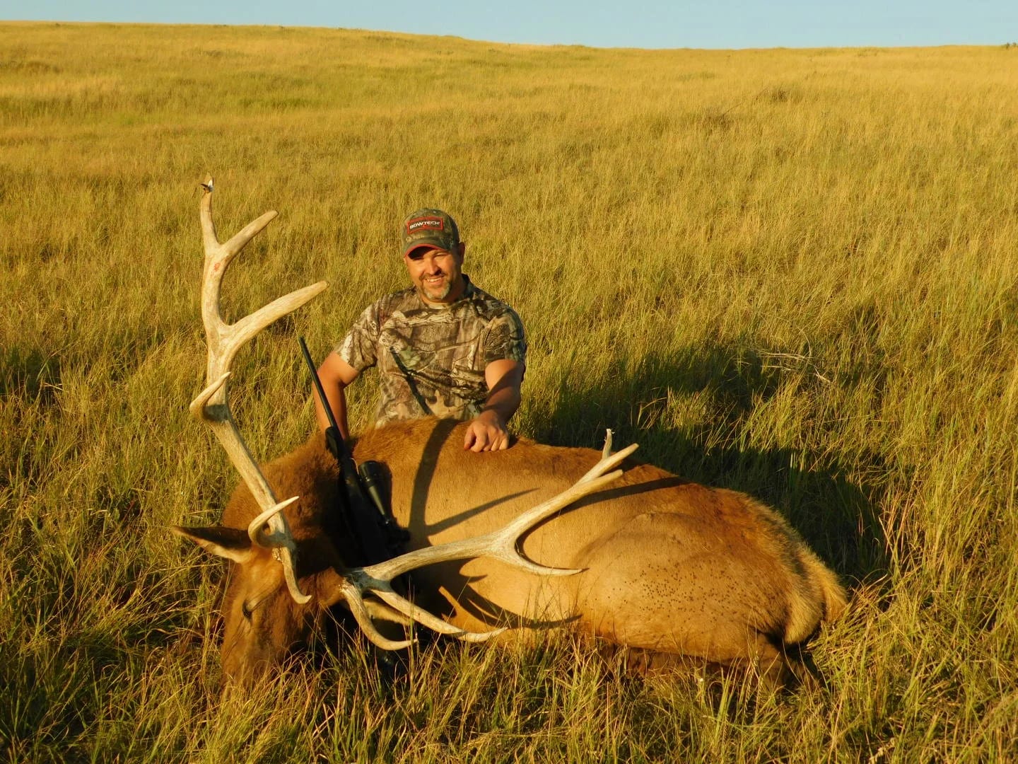 A hunter in camouflage poses with a harvested elk in a grassy field.