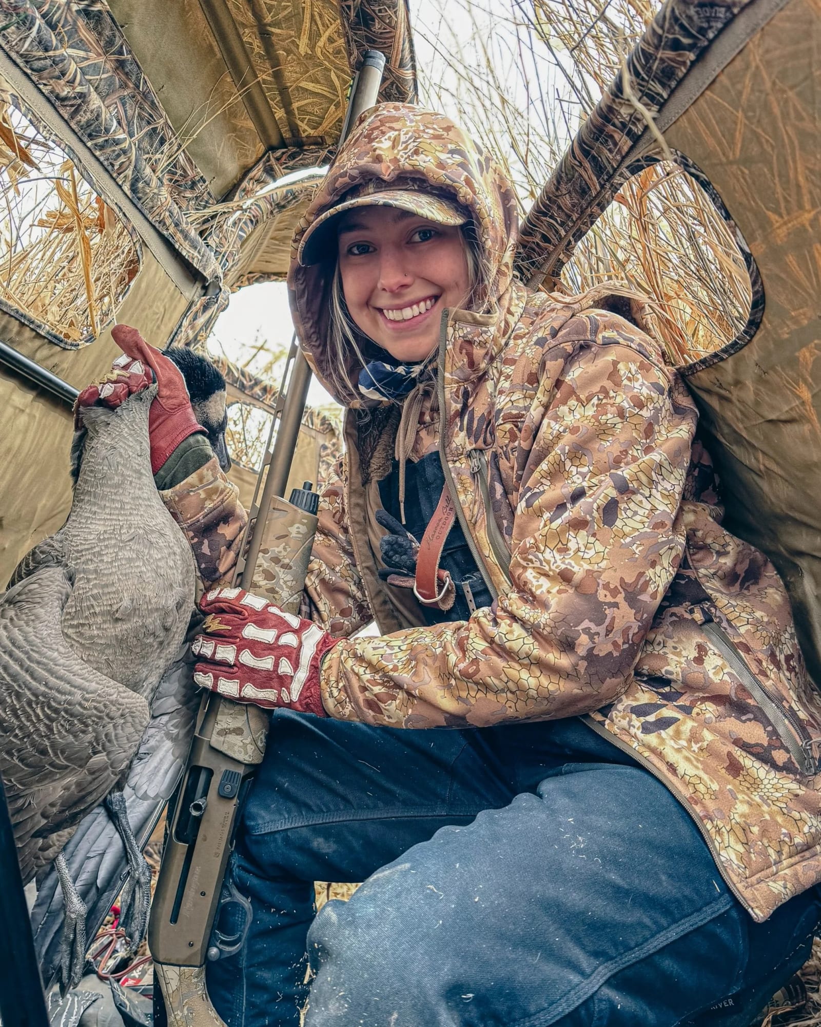A woman in camouflage clothing and a hat is sitting inside a hunting blind, smiling, holding a goose she has hunted, with a shotgun resting against her knee.