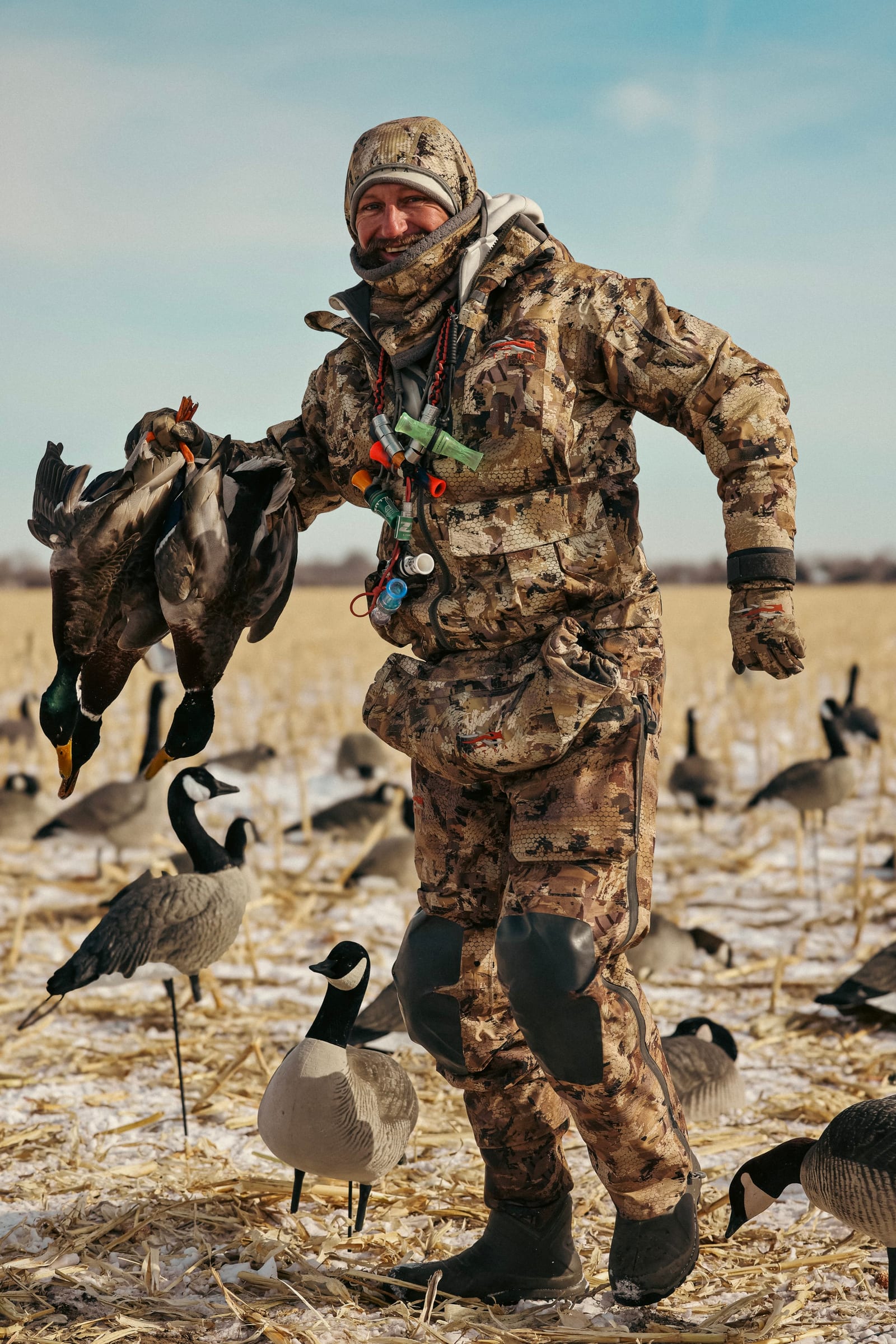 A man in camouflage hunting gear holding a string of dead ducks in a field with snow, surrounded by decoys of ducks.