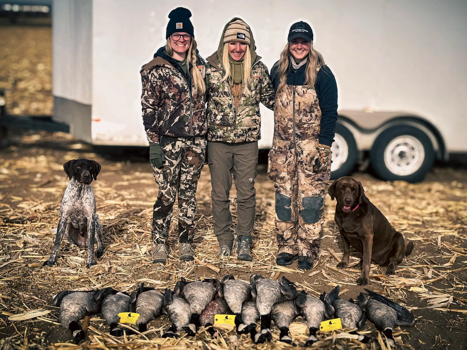 Three women in camouflage clothing standing outdoors with two dogs, in front of a trailer, with multiple ducks and goose on the ground.