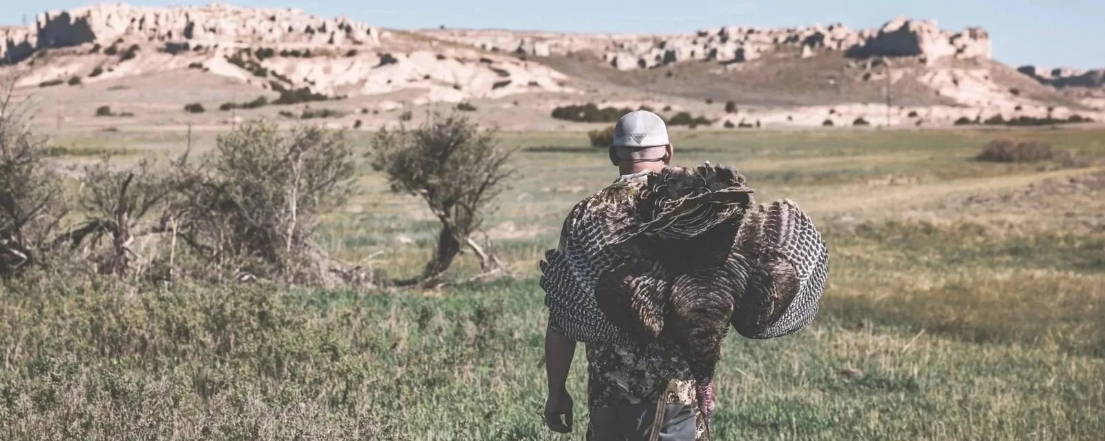 A person wearing camouflage clothing and a helmet, walking through a grassy field carrying a large turkey on their shoulders, with a backdrop of desert-like terrain and hills.