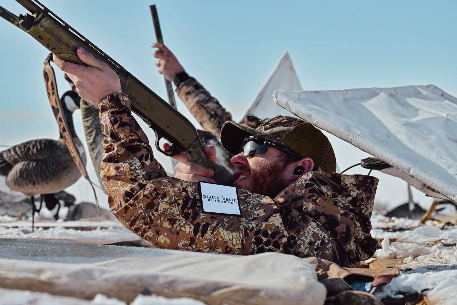 A man in camouflage clothing and sunglasses lying on snow, firing a rifle, with a large white flag and wildlife decoys in the background.