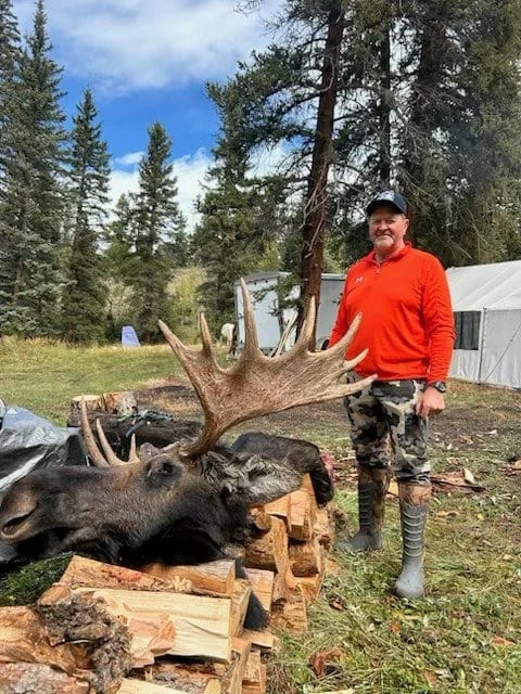 A man poses with a harvested moose lying on a pile of logs.