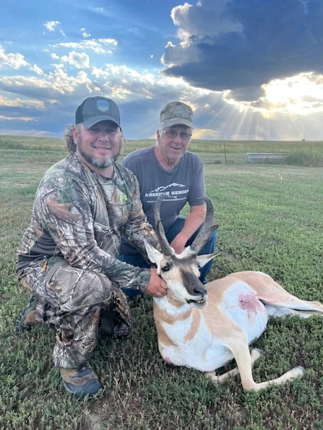 Two men pose with a harvested pronghorn in a grassy field under a cloudy sky.