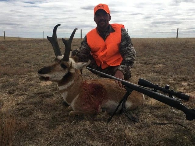 A hunter in an orange vest and hat poses with a harvested pronghorn and a rifle in a dry, open field.