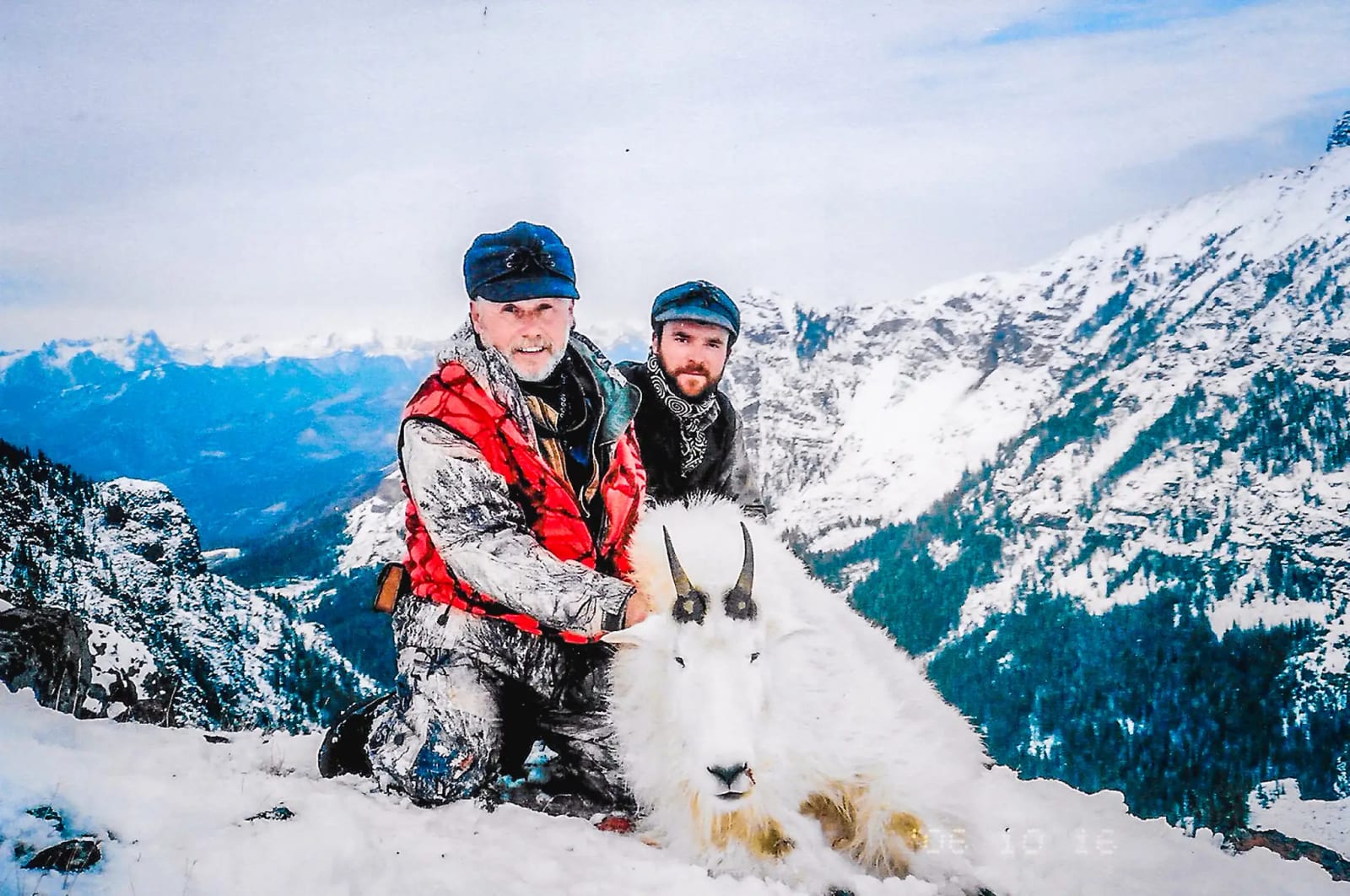 Two men pose with a harvested mountain goat in a snowy mountain landscape.