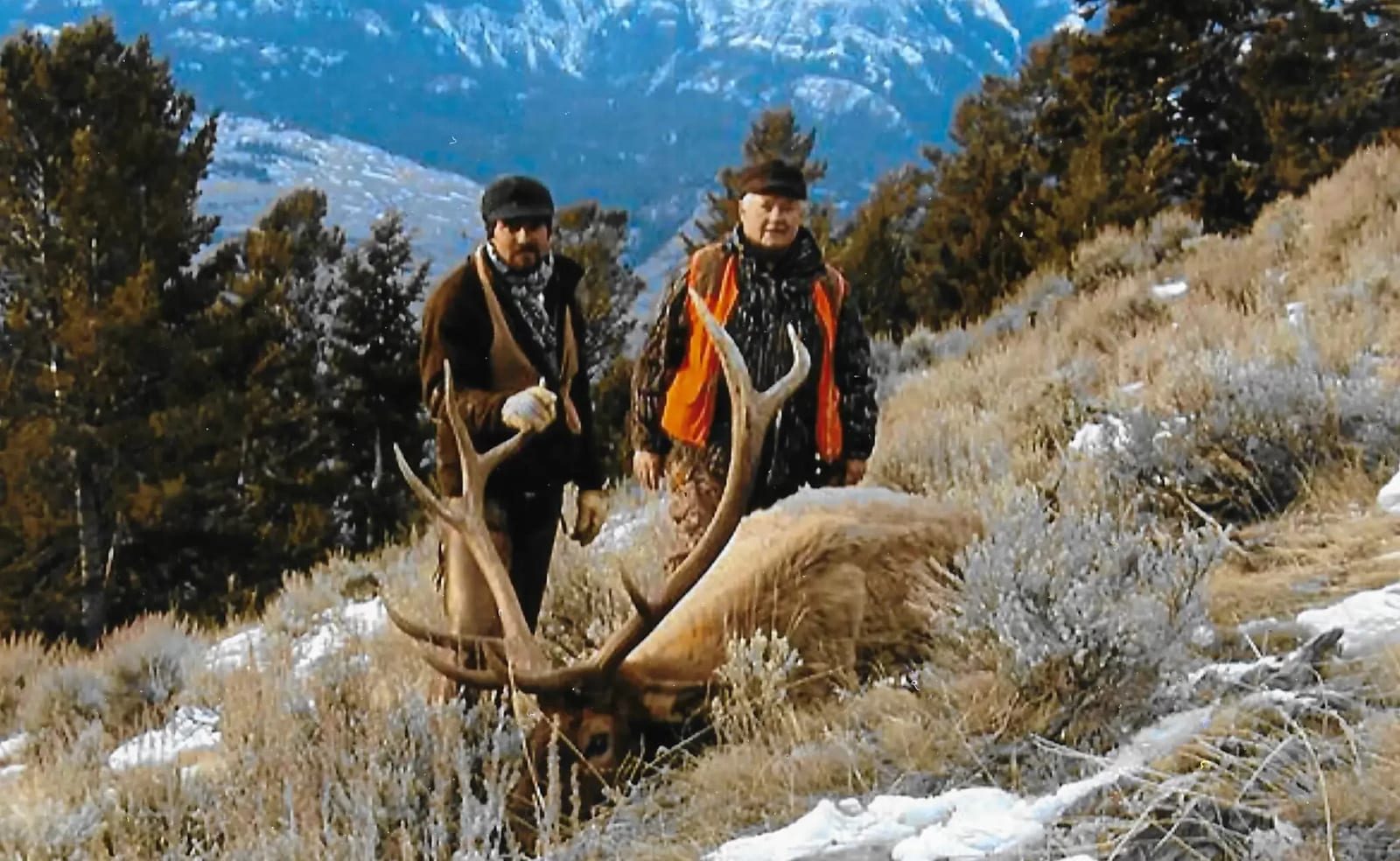 Two hunters pose with a harvested elk in a snowy, mountainous landscape.