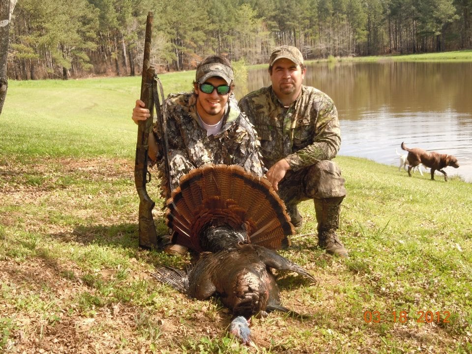 Two hunters pose with a harvested wild turkey and a shotgun next to a pond with a dog.