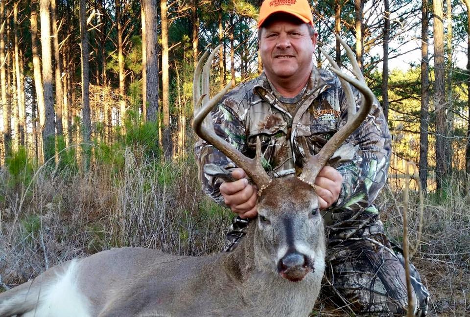 A man in camouflage and an orange hat poses with a harvested whitetail deer in a forest.