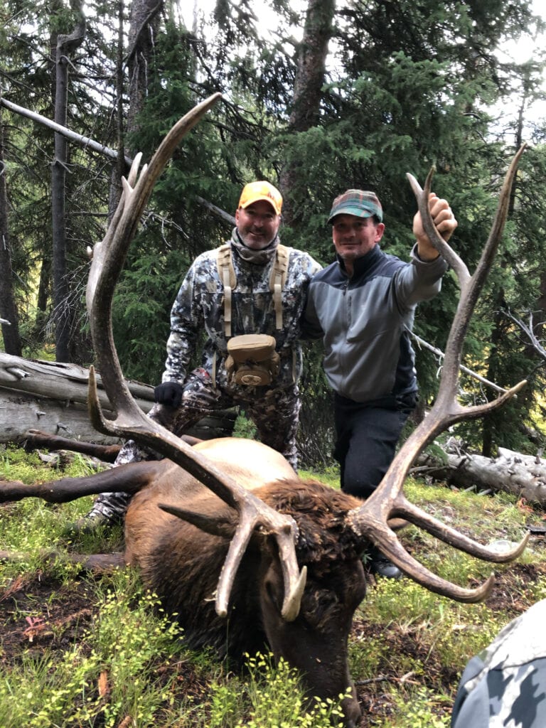 Two hunters pose with a harvested elk in a forest.