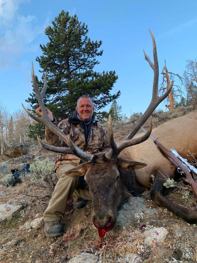 A hunter in camouflage poses with a harvested elk in a mountainous, forested area.