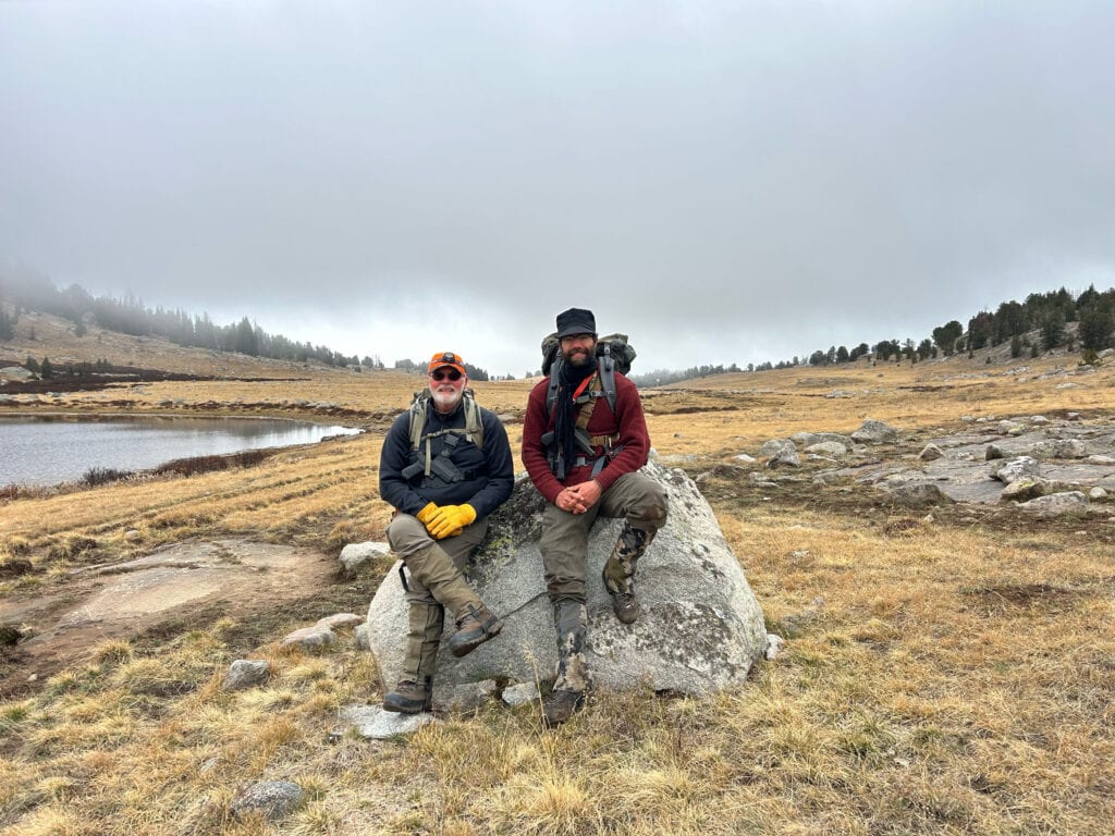 Two men in hunting gear sit on a large rock in a grassy field next to a lake under a cloudy sky.