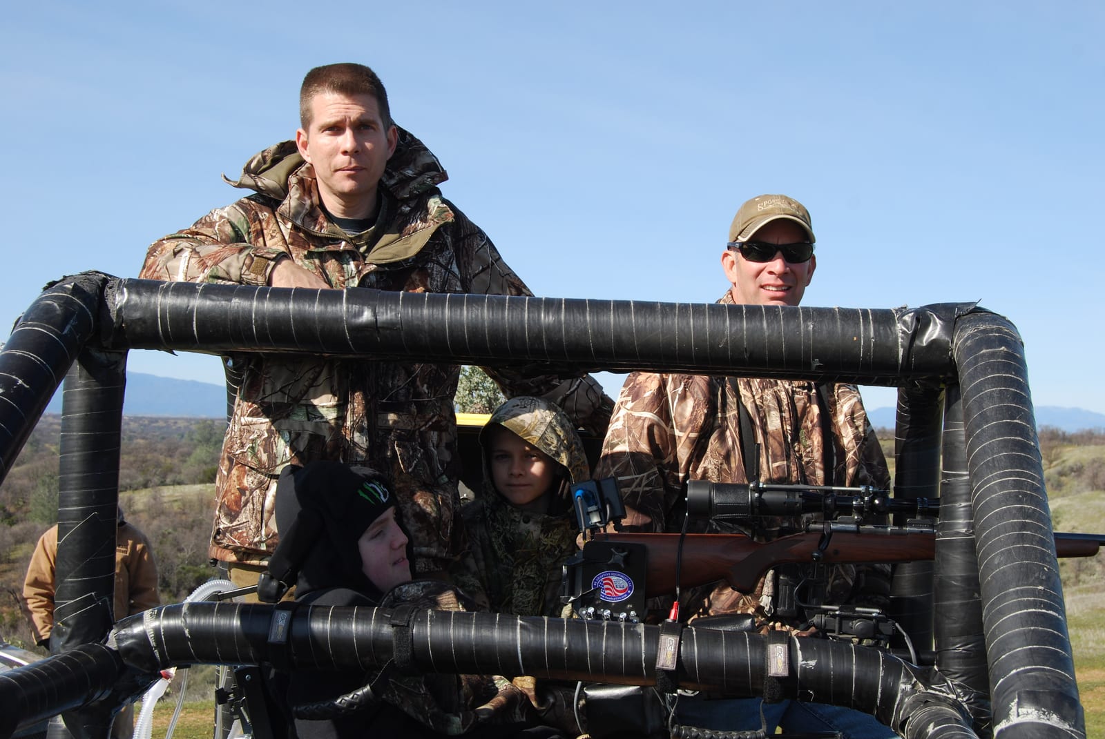 Several hunters in camouflage clothing are in a hunting vehicle in an outdoor setting.