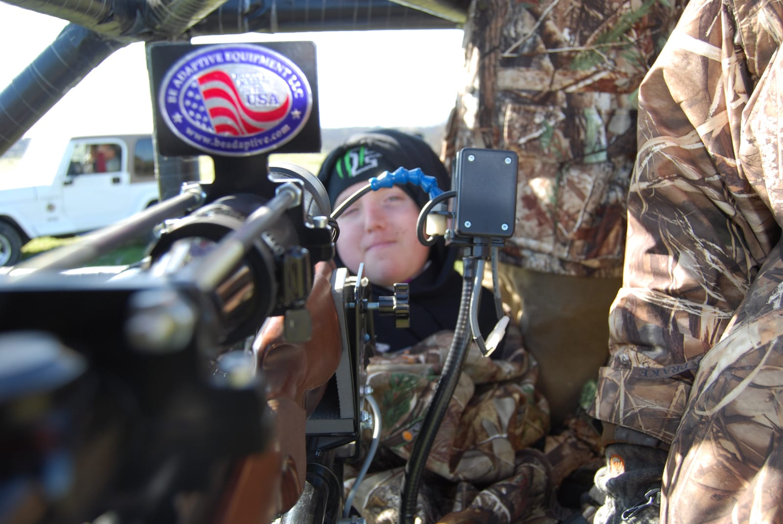 A person in camouflage operates adaptive hunting equipment from inside a vehicle or blind, with a white Jeep in the background.