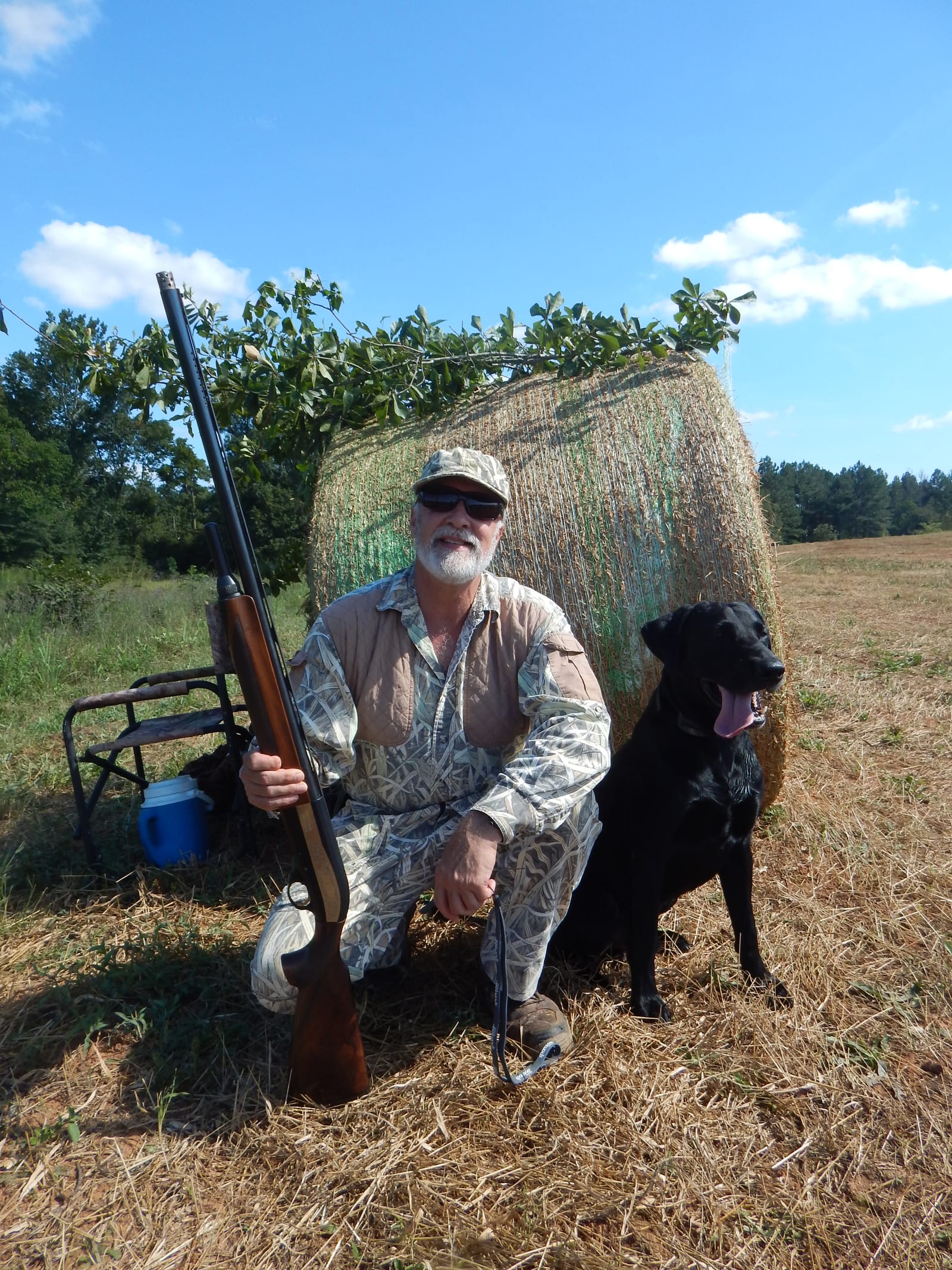 A man in camouflage kneels with a shotgun and a black dog in a field next to a hay bale.