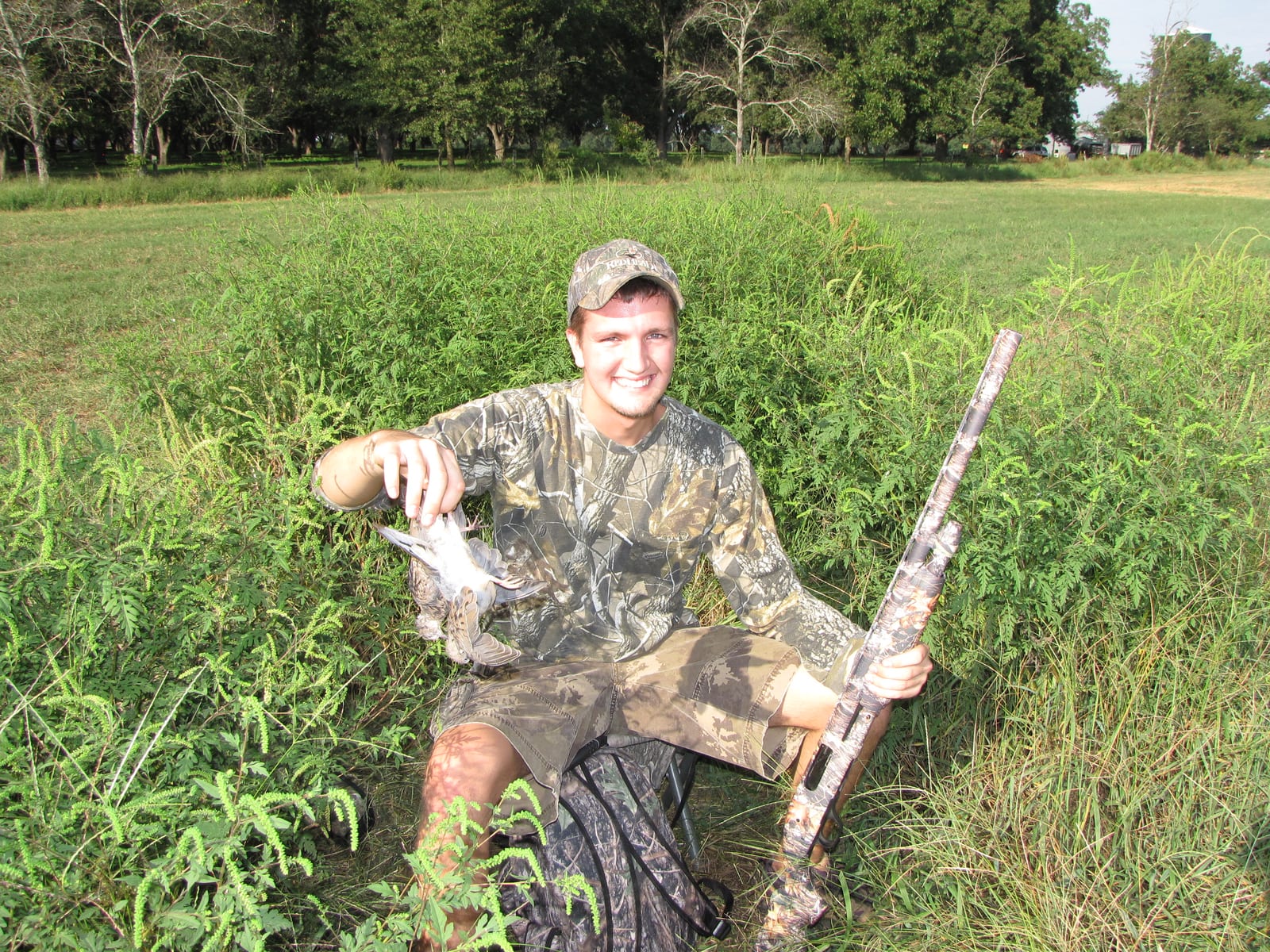 A hunter in camouflage holds a harvested mourning dove and a shotgun in a grassy field.