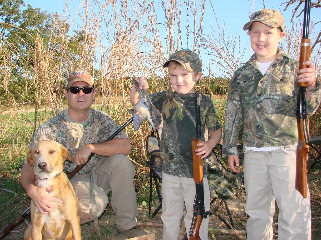 A man, two boys, and a dog pose with shotguns and a harvested mourning dove in a field.