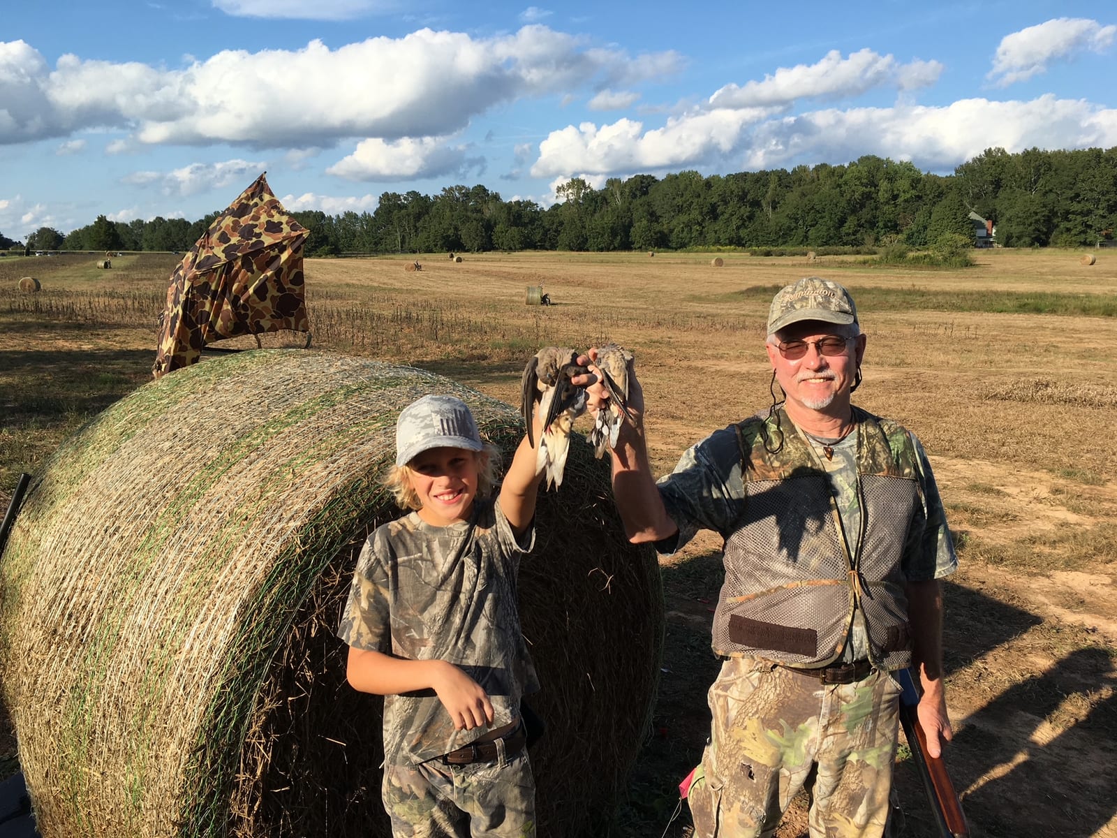 A man and a boy in camouflage clothing pose with harvested mourning doves in a field with hay bales.