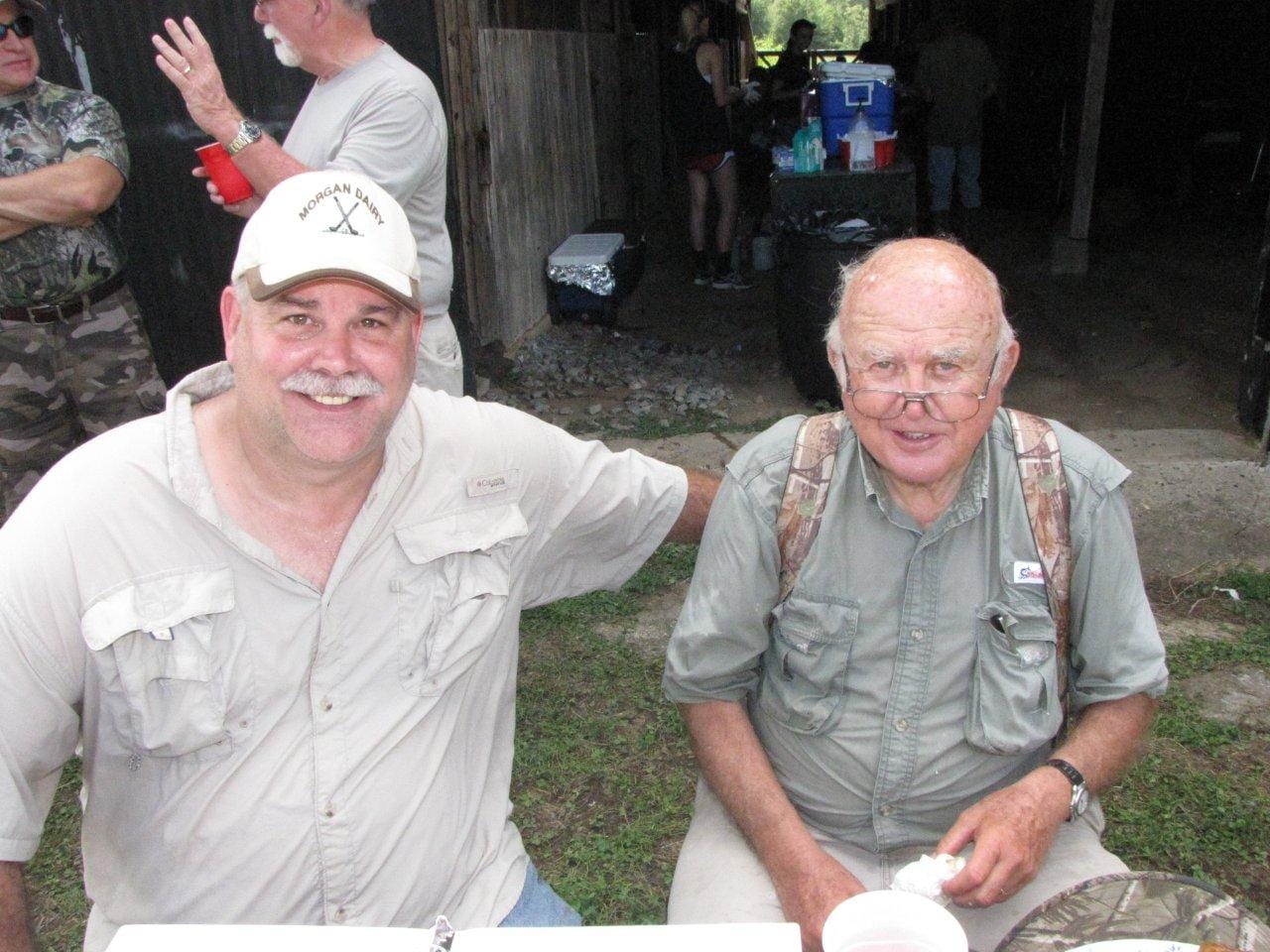 Two men smile at the camera, one wearing a white cap and the other wearing glasses and a camouflage-patterned vest.