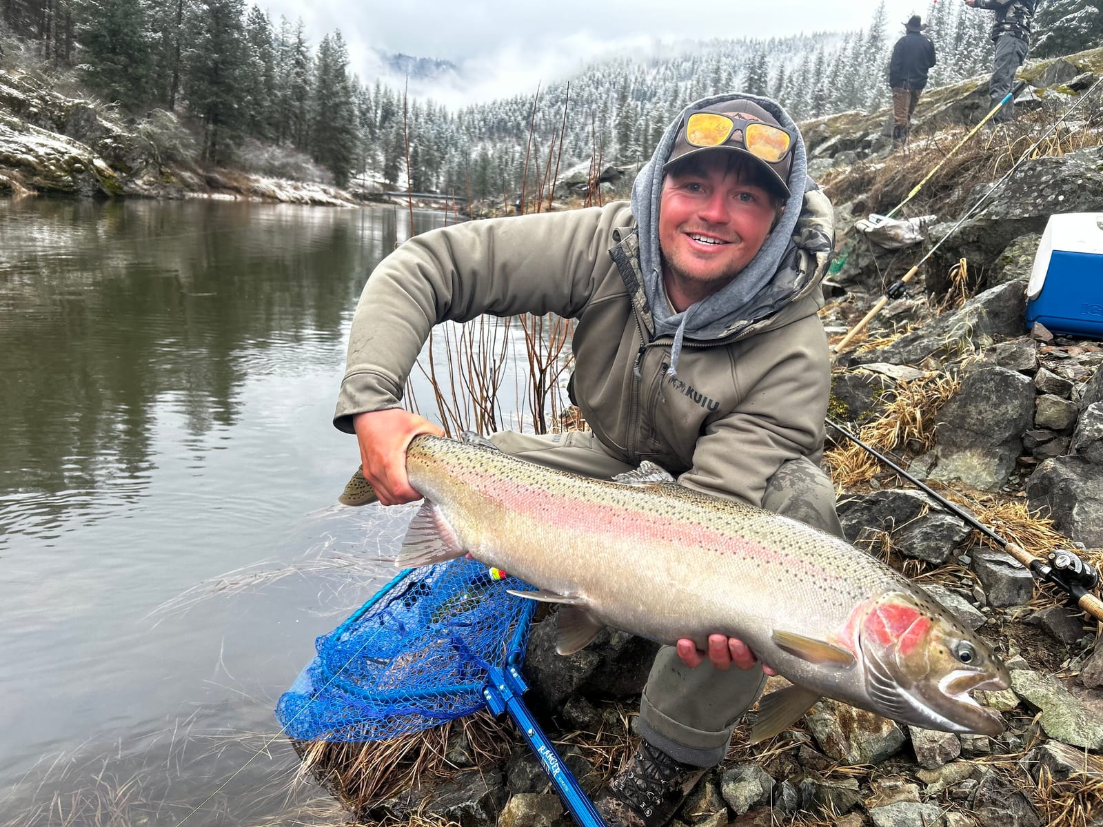 A man in a hooded jacket and sunglasses holds a large rainbow trout by a river with snowy trees in the background.