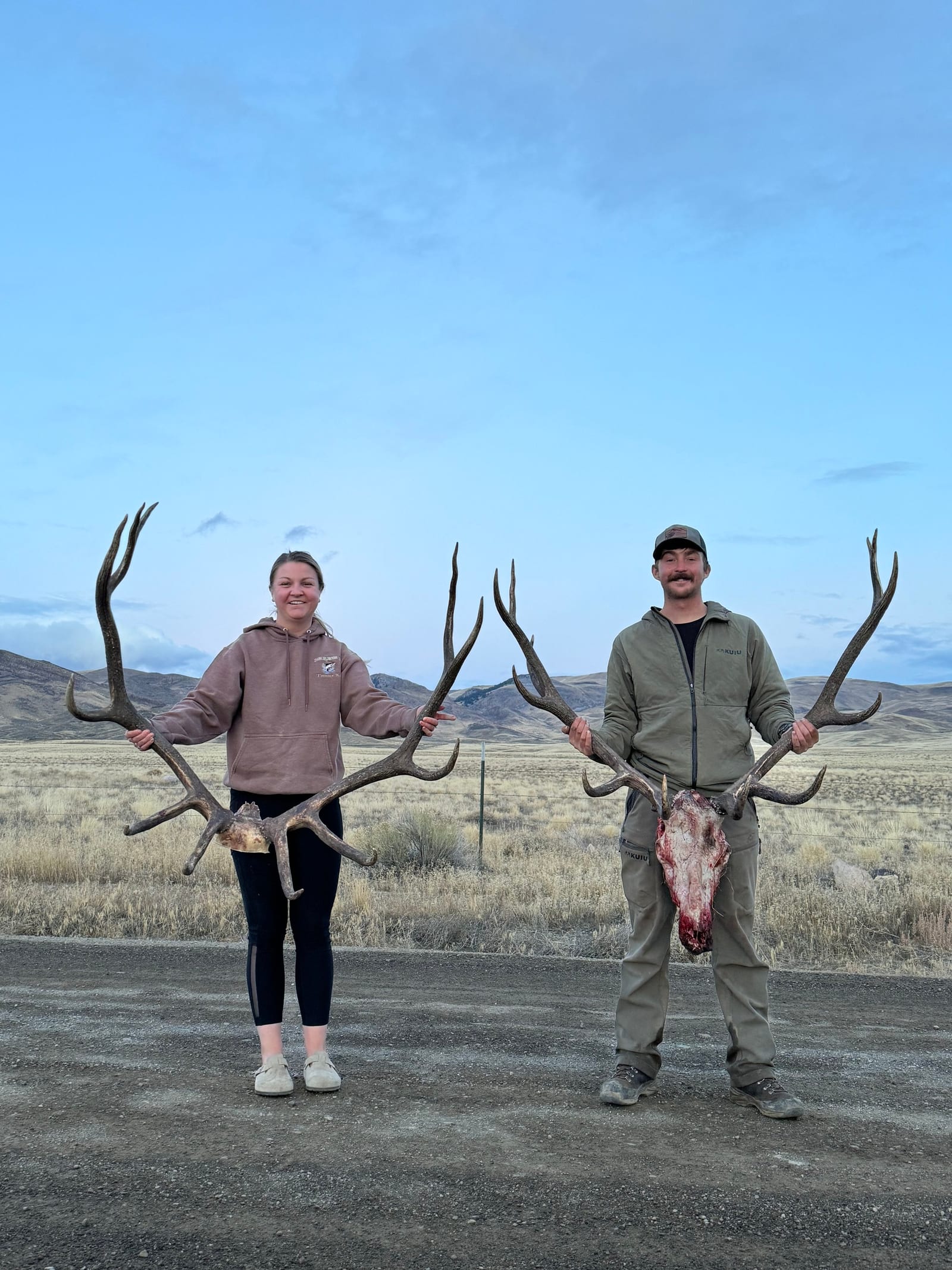 Two hunters pose with harvested elk antlers and a skull in a field.