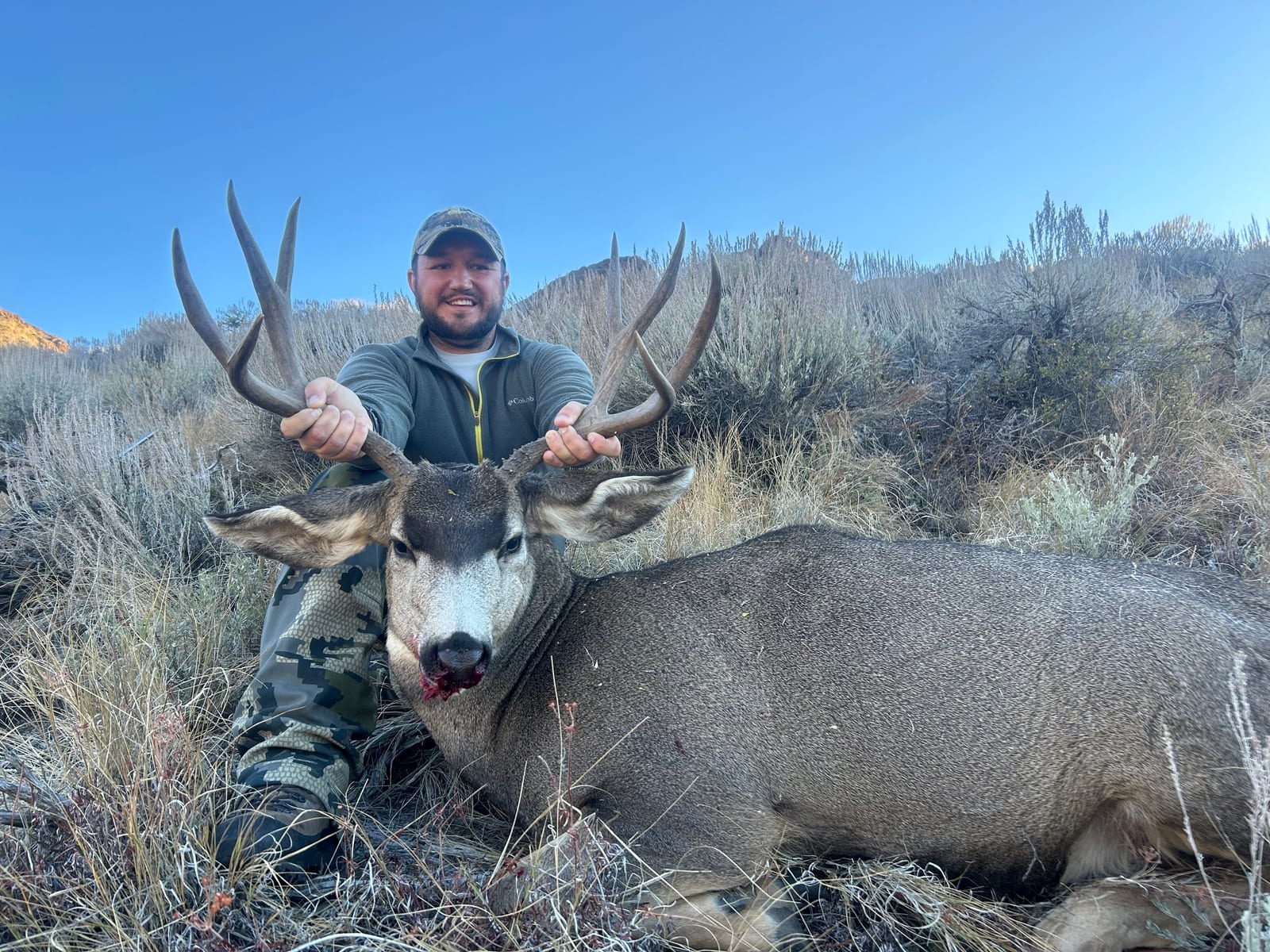 A hunter poses with a harvested mule deer in a field.