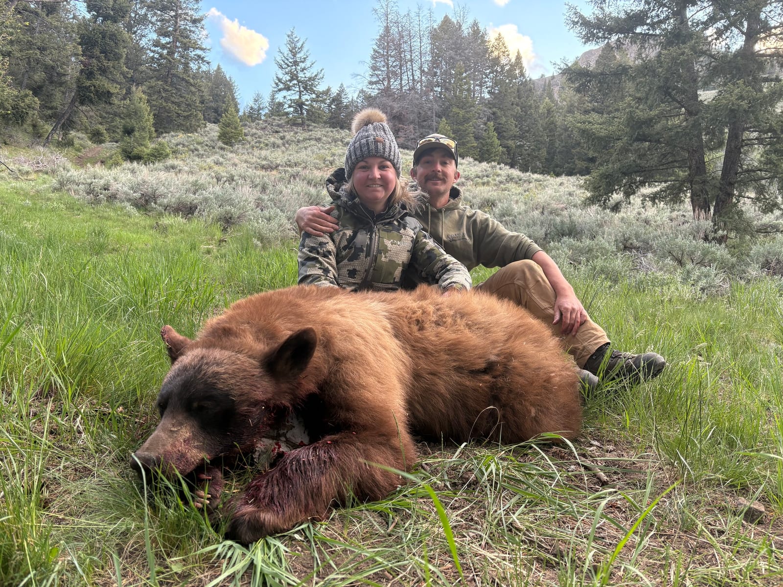 Two hunters pose with a harvested black bear in a grassy, wooded area.
