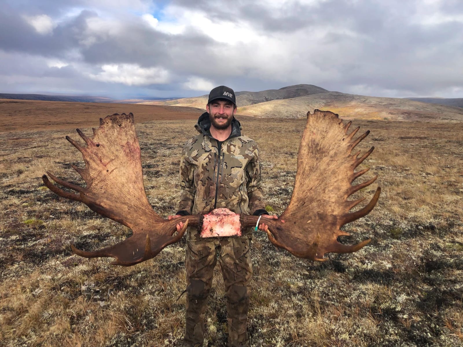 A hunter poses with large moose antlers and a piece of meat in a vast, open landscape.