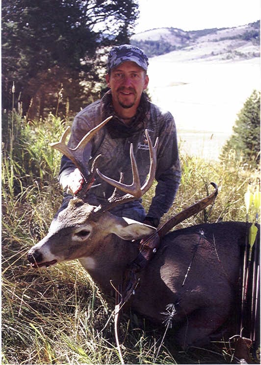 A hunter in camouflage poses with a harvested whitetail deer in a grassy, wooded area.