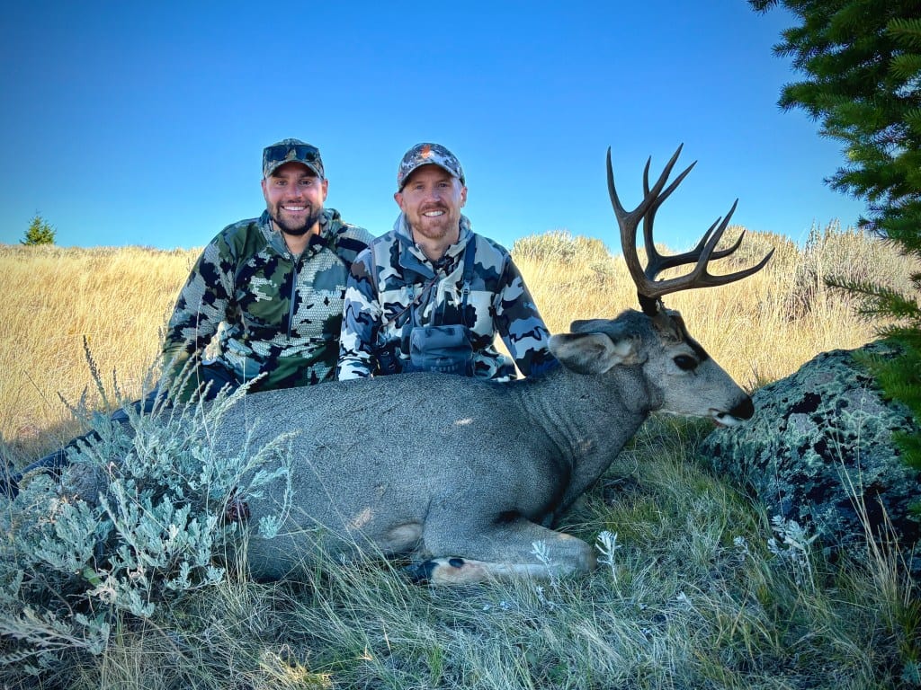 Two hunters pose with a harvested mule deer in a grassy field.