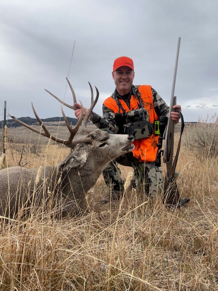 A hunter in an orange vest and hat kneels beside a harvested mule deer, holding its antlers, with a rifle leaning nearby in a grassy field.