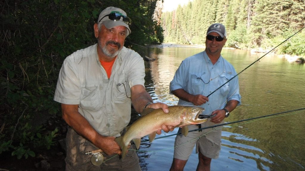 Two men stand in a river, one holding a large fish, with trees lining the banks.