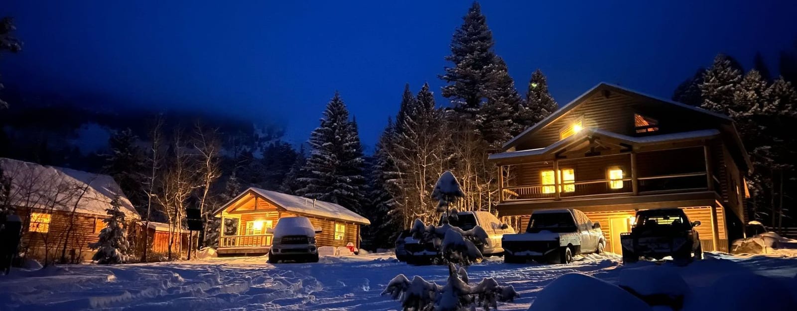 Several cabins and vehicles are covered in snow at night, illuminated by lights from the buildings.