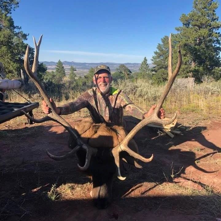 A man in camouflage clothing holding large antlers and sitting behind a dead elk in a natural outdoor setting with trees and mountains in the background.