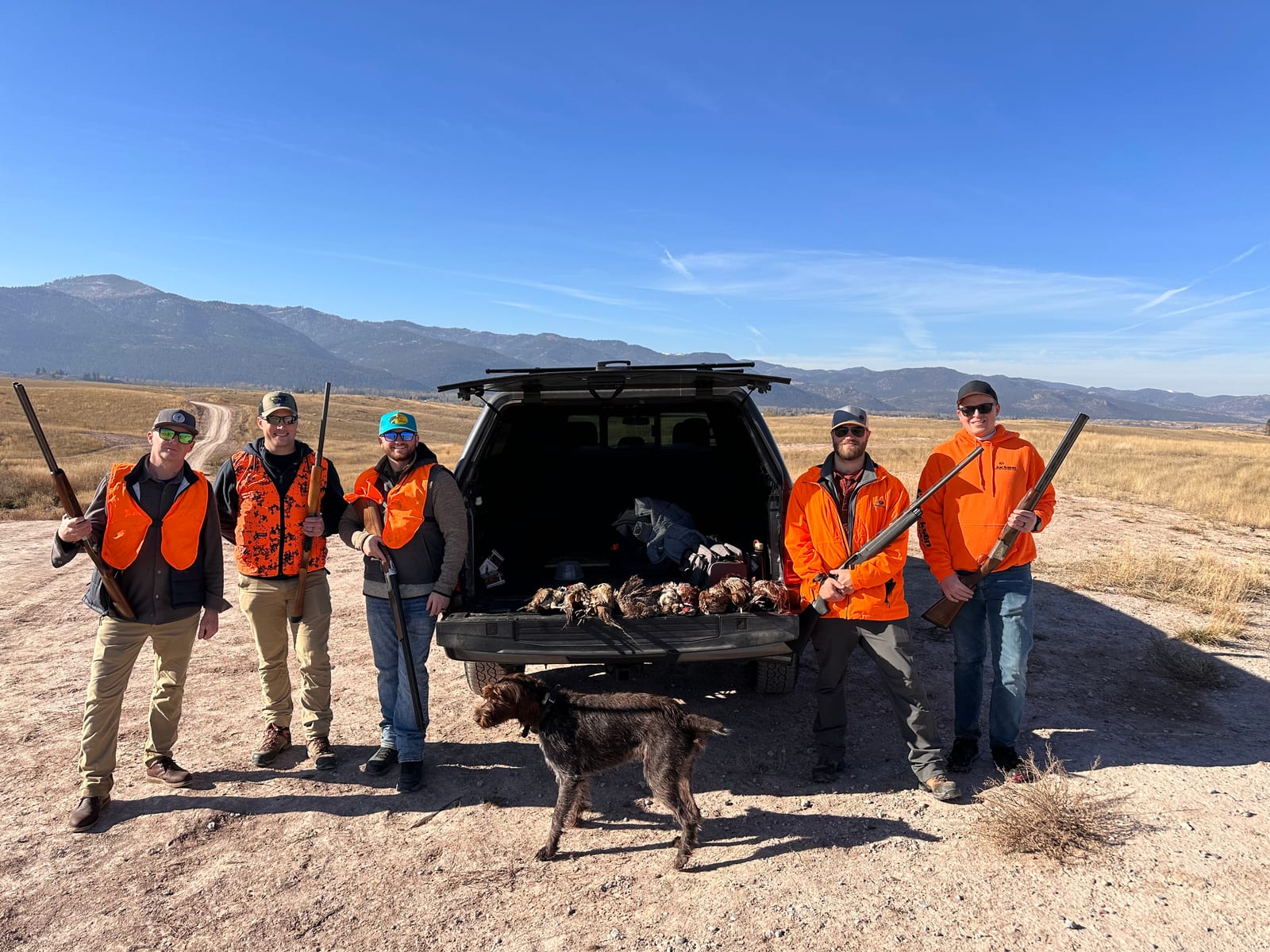 Five hunters and a dog pose with harvested pheasants next to a truck in a field.